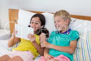 Smiling siblings using digital tablet on bed in bedroom