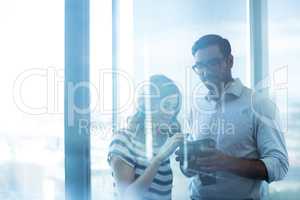 Business couple holding digital camera against glass window in office