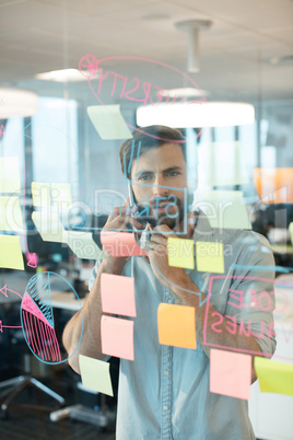 Businessman using mobile phone while looking at adhesive notes an graphs on glass