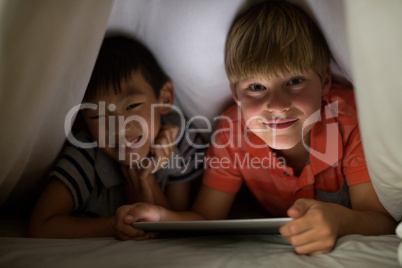 Portrait of siblings under bed sheet using digital tablet on bed
