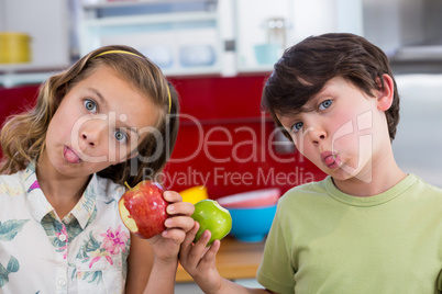 Siblings holding apple and pulling funny faces in kitchen