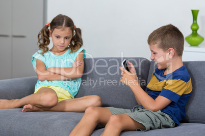 Upset girl sitting on sofa while boy using mobile phone in living room