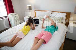 Siblings using mobile phone and laptop on bed