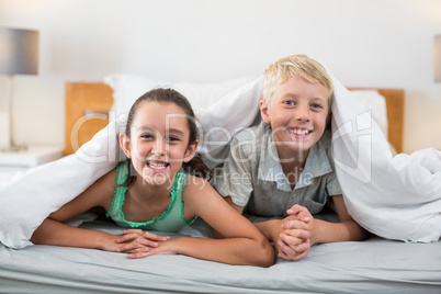 Smiling siblings lying under bed sheet in bedroom