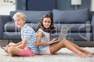 Siblings sitting back to back and using digital tablet and laptop