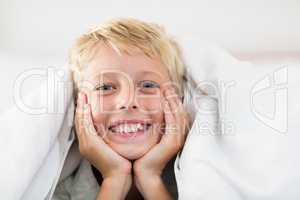 Portrait of smiling boy lying under bed sheet in bedroom