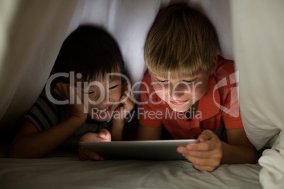 Siblings under bed sheet using digital tablet on bed