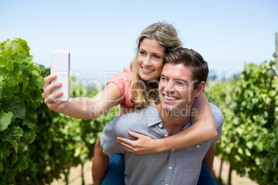 Happy couple taking selfie while piggybacking at vineyard against sky