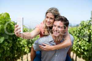 Happy couple taking selfie while piggybacking at vineyard against sky