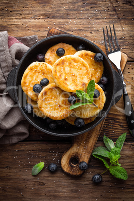 Delicious cottage cheese pancakes or syrniki with fresh blueberry in cast-iron pan on dark wooden rustic background, above view. Tasty breakfast.