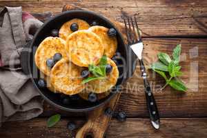 Delicious cottage cheese pancakes or syrniki with fresh blueberry in cast-iron pan on dark wooden rustic background, above view. Tasty breakfast.