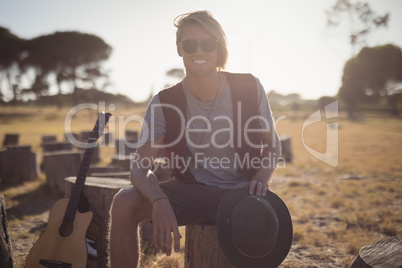 Portrait of smiling young man sitting by guitar on tree stump