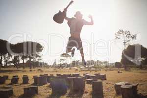 Young man holding guitar while jumping on tree stump