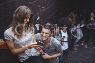 Happy friends using mobile phones while standing by wall