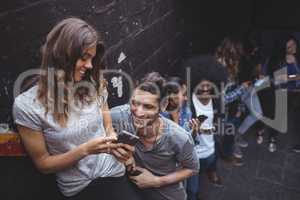 Happy friends using mobile phones while standing by wall