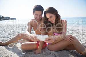 Smiling couple taking selfie while sitting at beach