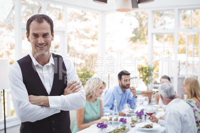 Smiling waiter standing with arms crossed while friends dining in background