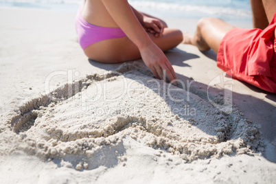 Mid section of couple sitting by heart shape carved on sand at beach