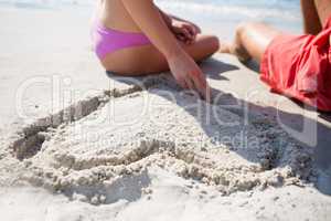 Mid section of couple sitting by heart shape carved on sand at beach