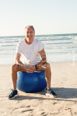 Portrait of smiling senior man sitting on exercise ball against sea