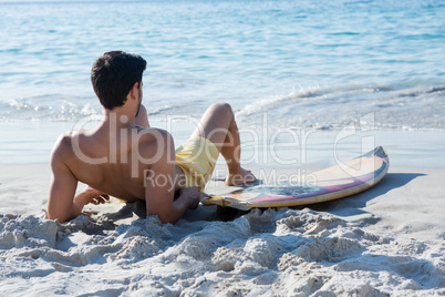 Full length of shirtless man reclining by surfboard at beach