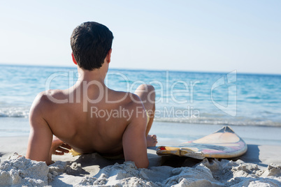 Rear view of shirtless man reclining by surfboard at beach