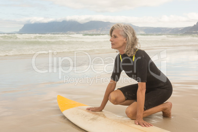 Senior woman kneeling by surfboard on shore