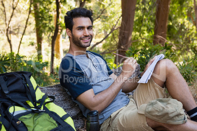 Smiling man writing on notepad while resting on tree trunk
