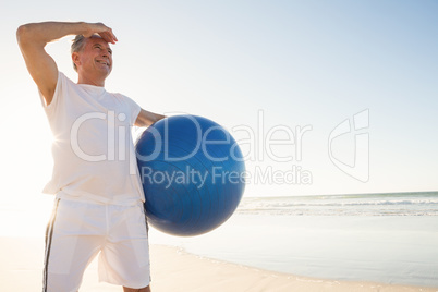 Senior man looking away while holding exercise ball at beach