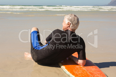 Rear view of senior man in wetsuit sitting by surfboard