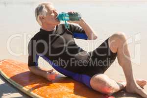 Senior man drinking water while sitting by surfboard