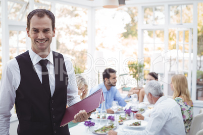 Smiling waiter holding menu while friends dining in background