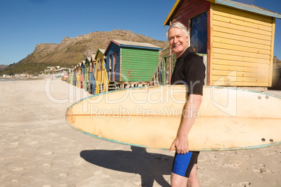 Portrait of smiling man carrying surfboard standing against beach hut