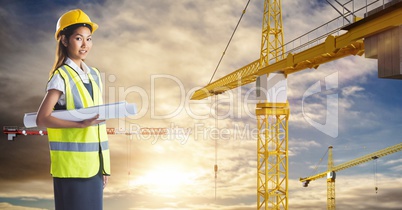 Female architect holding blueprint by cranes against sky
