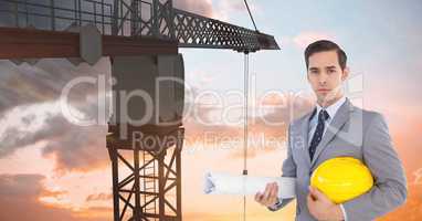Architect holding hardhat and blueprint in front of crane