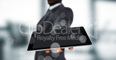 Business man mid section holding out tablet behind bokeh against blurry grey window