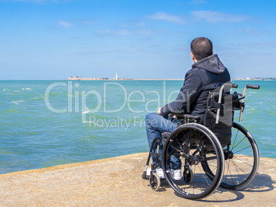 Disabled young man sitting in a wheelchair and looks at the sea