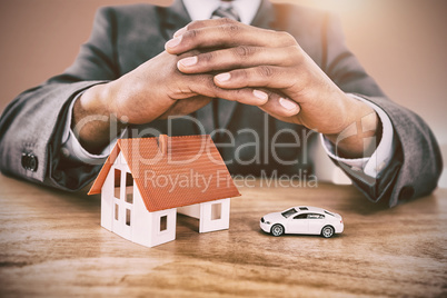 Composite image of businessman protecting house model and car with hands on table