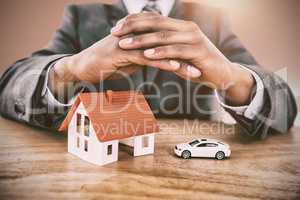 Composite image of businessman protecting house model and car with hands on table