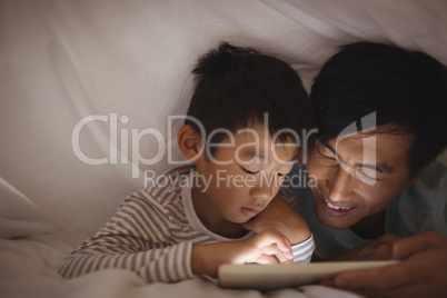 Father and son using digital tablet under blanket in bedroom