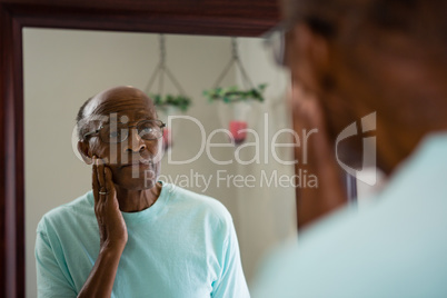 Concerned senior man rubbing cheek while looking into mirror