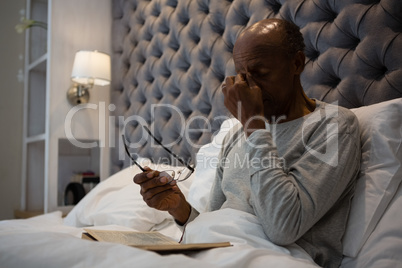 Tired senior man massaging eyes while holding eyeglasses on bed