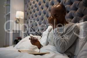 Tired senior man massaging eyes while holding eyeglasses on bed