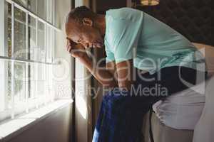 Side view of serious senior man sitting on bed by window