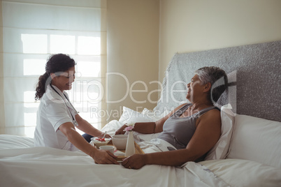 Female doctor serving breakfast to senior woman on bed