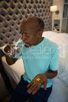 Senior man holding medicine while drinking water in bedroom