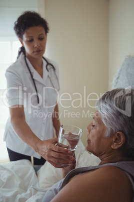 Female doctor giving sick senior woman a glass of water on bed