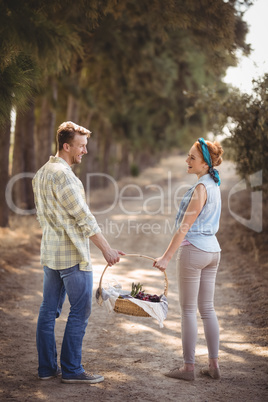 Cheerful young couple carrying basket on dirt road at farm