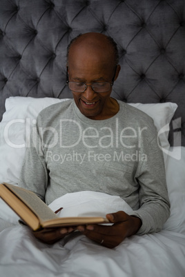 Smiling senior man reading book while sitting on bed