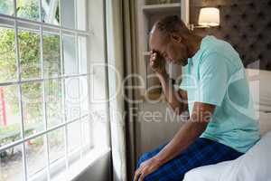 Side view of sad senior man sitting on bed by window at home